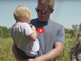 image of a farmer wearing a grey tshirt holding a child and a red video play button