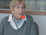 Image of woman with short hair and a grey sweater speaking with a video play icon in red