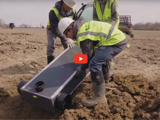 image of people moving items on top of dirt wearing neon green vests and a red video play button